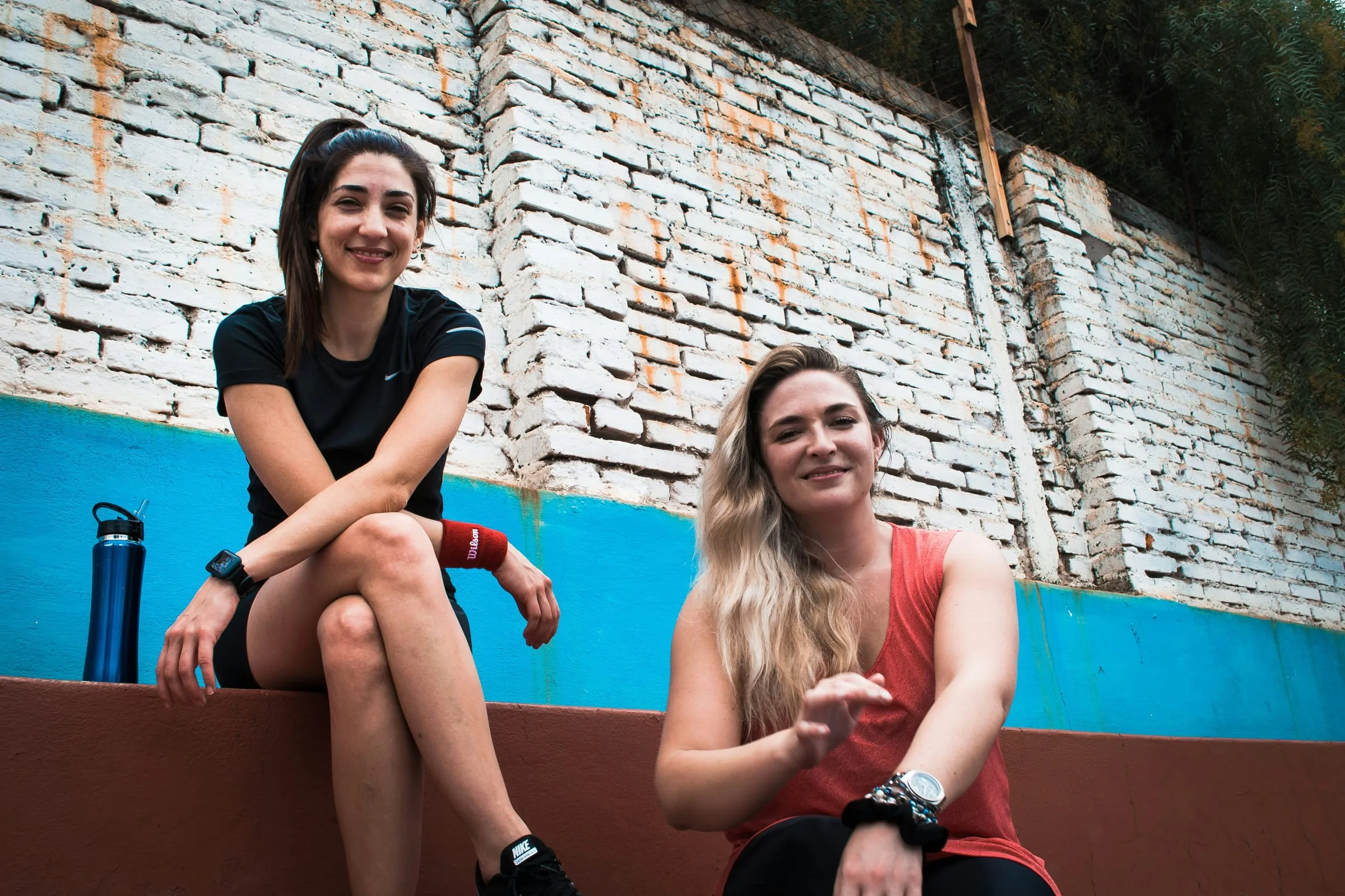 Two players resting on a courtside wall, smiling, a water bottle beside them