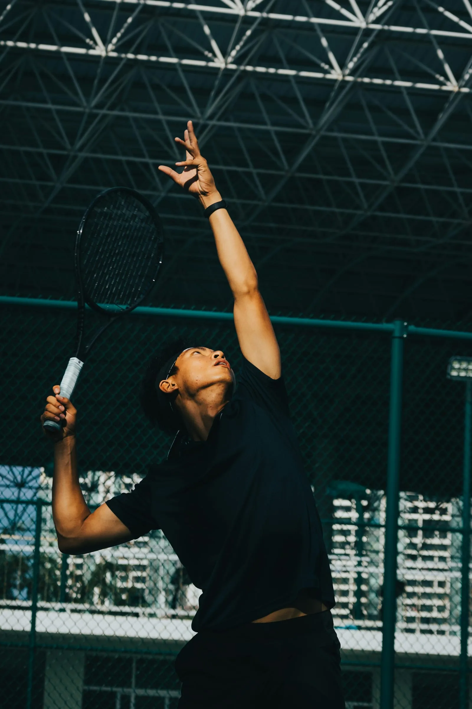 A tennis player reaching up to serve under a covered steel roof