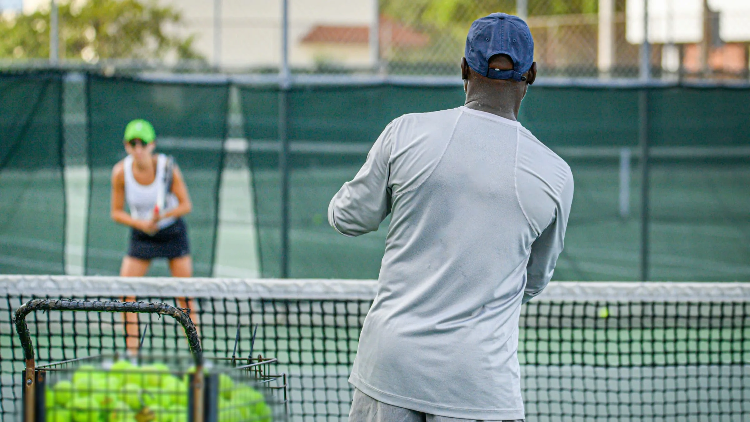 A tennis coach at the net rallying with a player, a basket of balls beside him