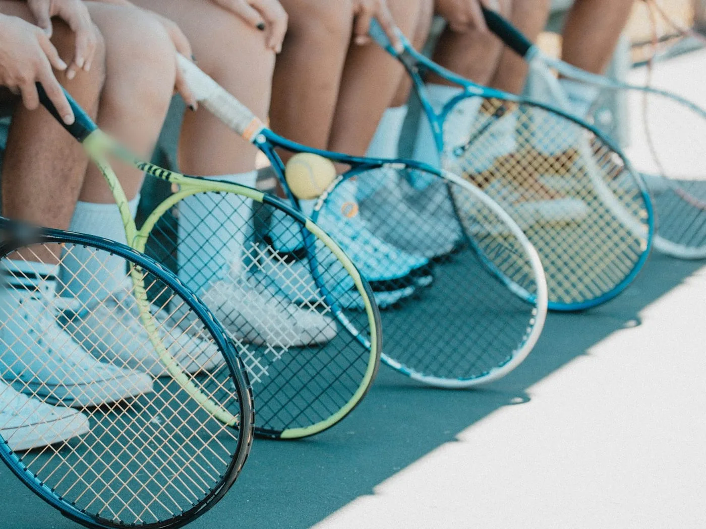 Young players sitting in a row on a courtside bench, racquets resting at their feet