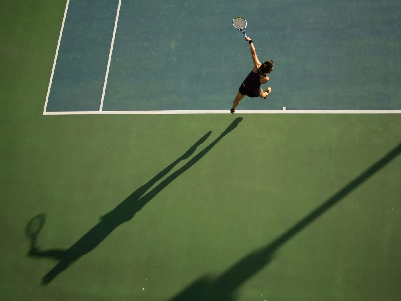 A tennis player serving, seen from above, her long shadow across the court