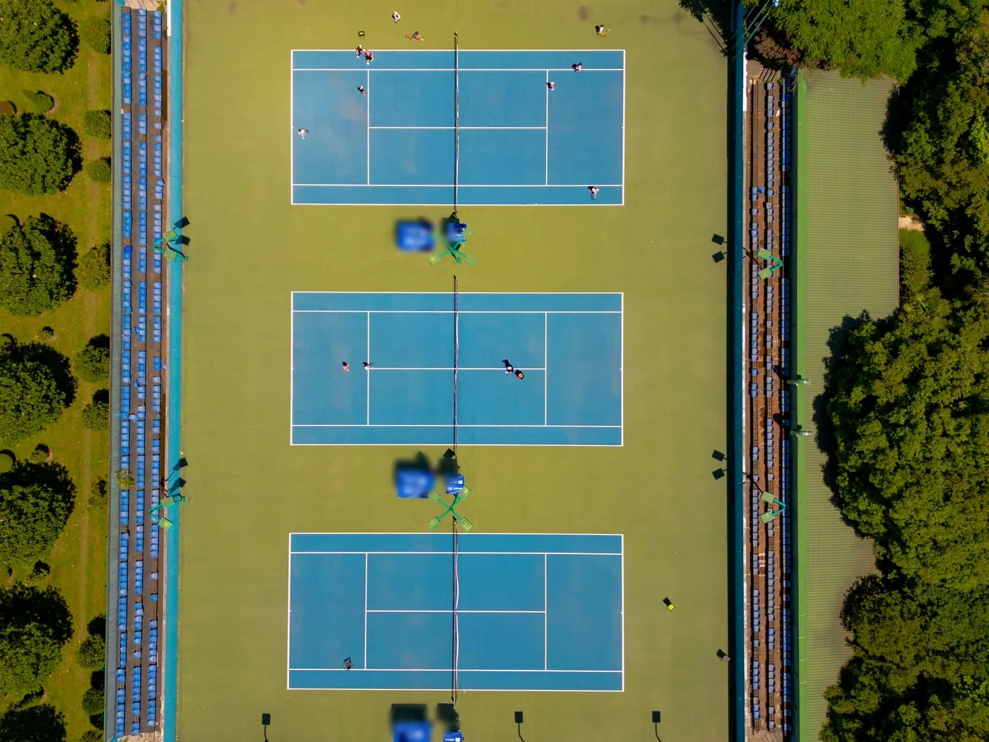 Three tennis courts and spectator stands among trees, seen from above